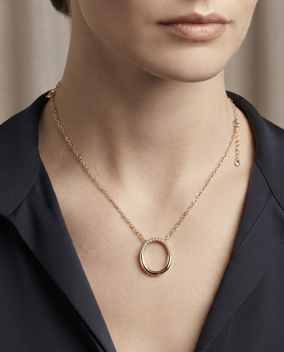 A close-up of a model wearing a delicate gold chain necklace with a double-ring pendant, one of which is set with small clear stones. The model is wearing a dark navy blue satin top against a soft beige background.